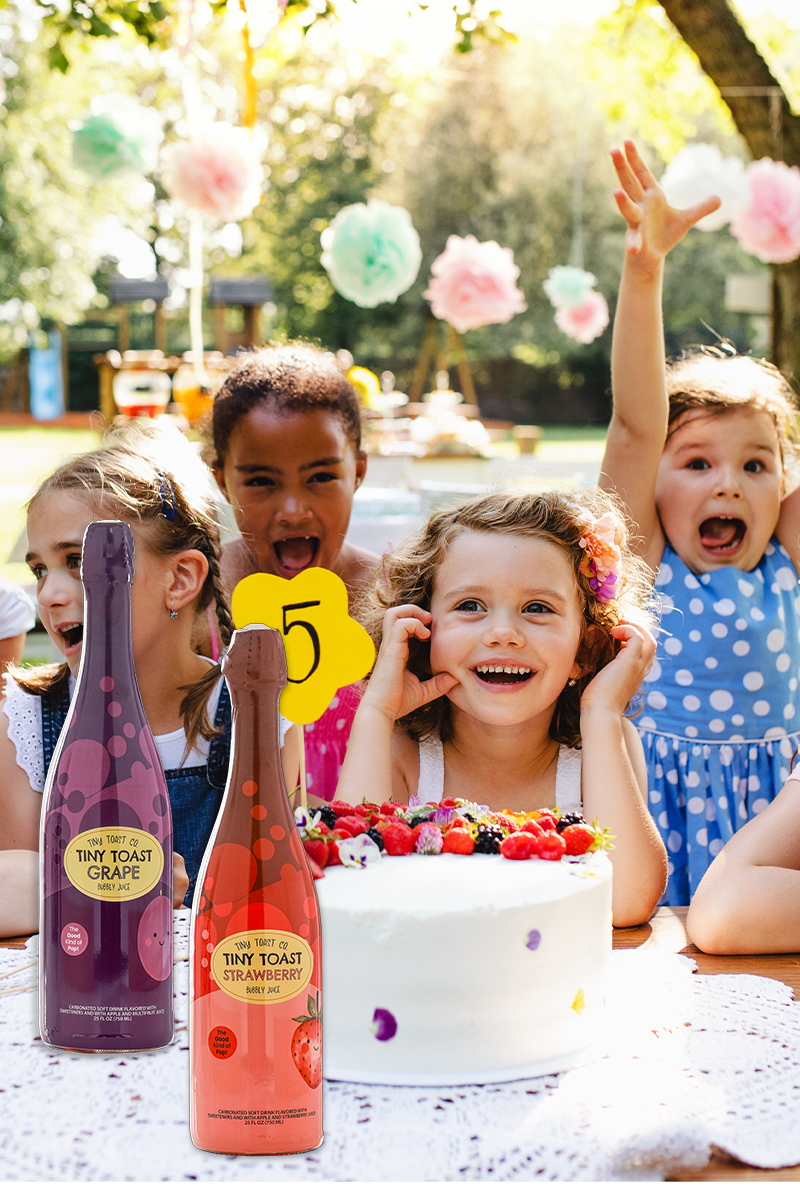 Three children sitting outdoors at a party with a birthday cake and two miniature beverage bottles in the foreground. The bottles are labeled 'TINY TOAST' and one has a purple label reading 'GRAPE' and the other has an orange label reading 'CIDER'.