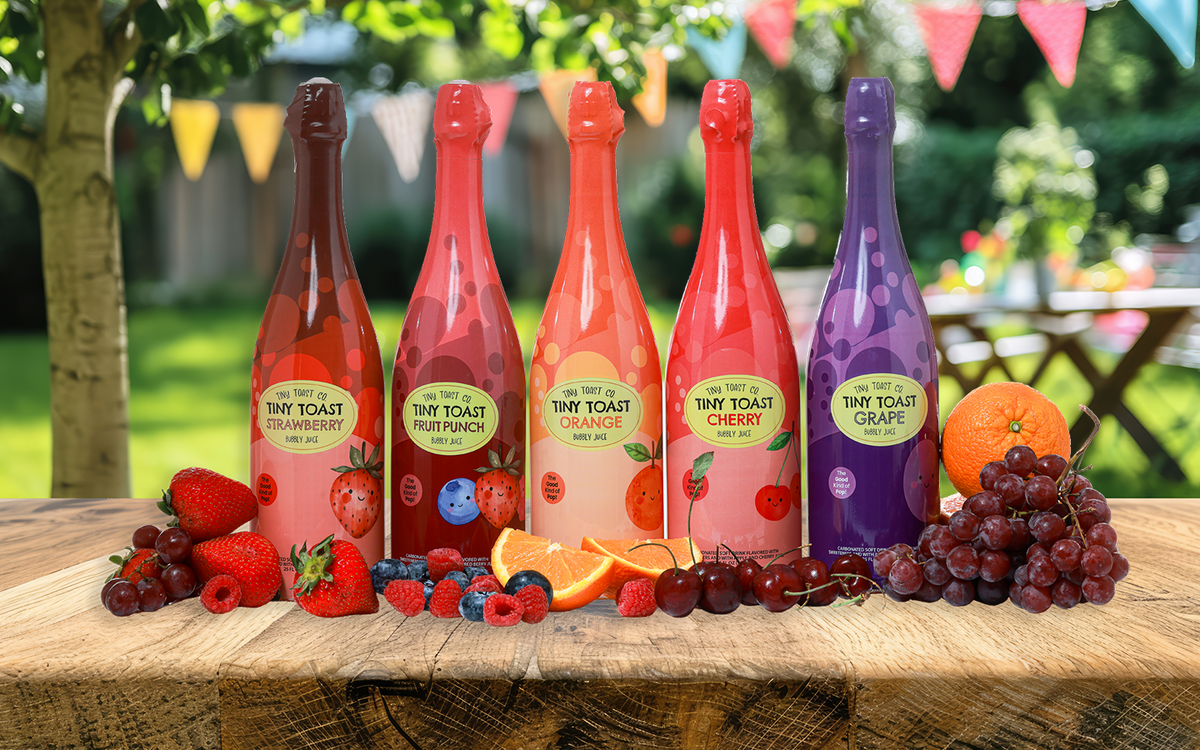 Six sparkling wine bottles from Tiny Toast lined up on a wooden table outdoors, surrounded by fresh fruit including strawberries, grapes, cherries, blueberries, and an orange. The background shows a blurred garden setting with colorful bunting.