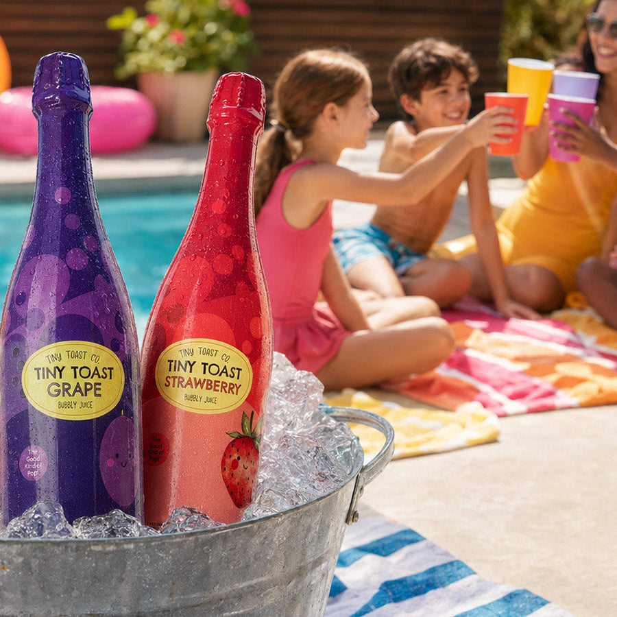 Three sparkling beverage bottles labeled Thin Toast Orange, Thin Toast Grape, and Thin Toast Strawberry in a metal ice bucket in front of a poolside scene with children and a woman clinking cups.
