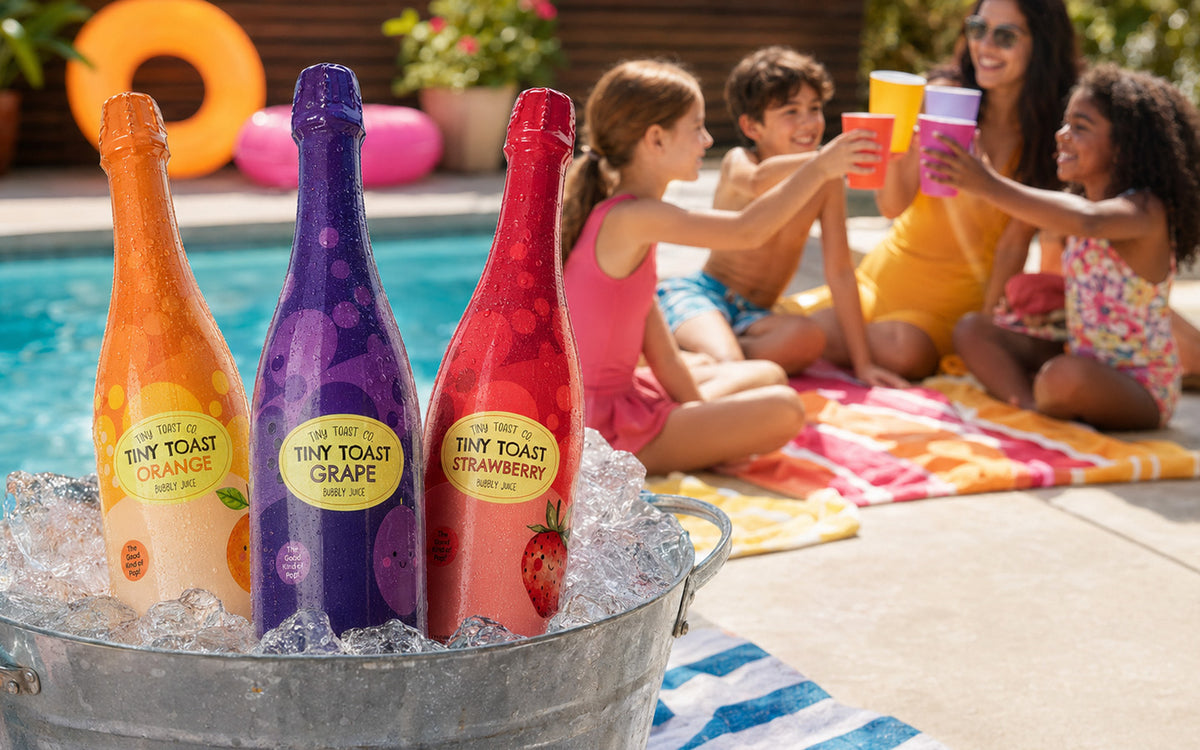 Three bottles of Tiny Toast Sparkling Juice by the pool at a kids party with the kids and parent toasting.