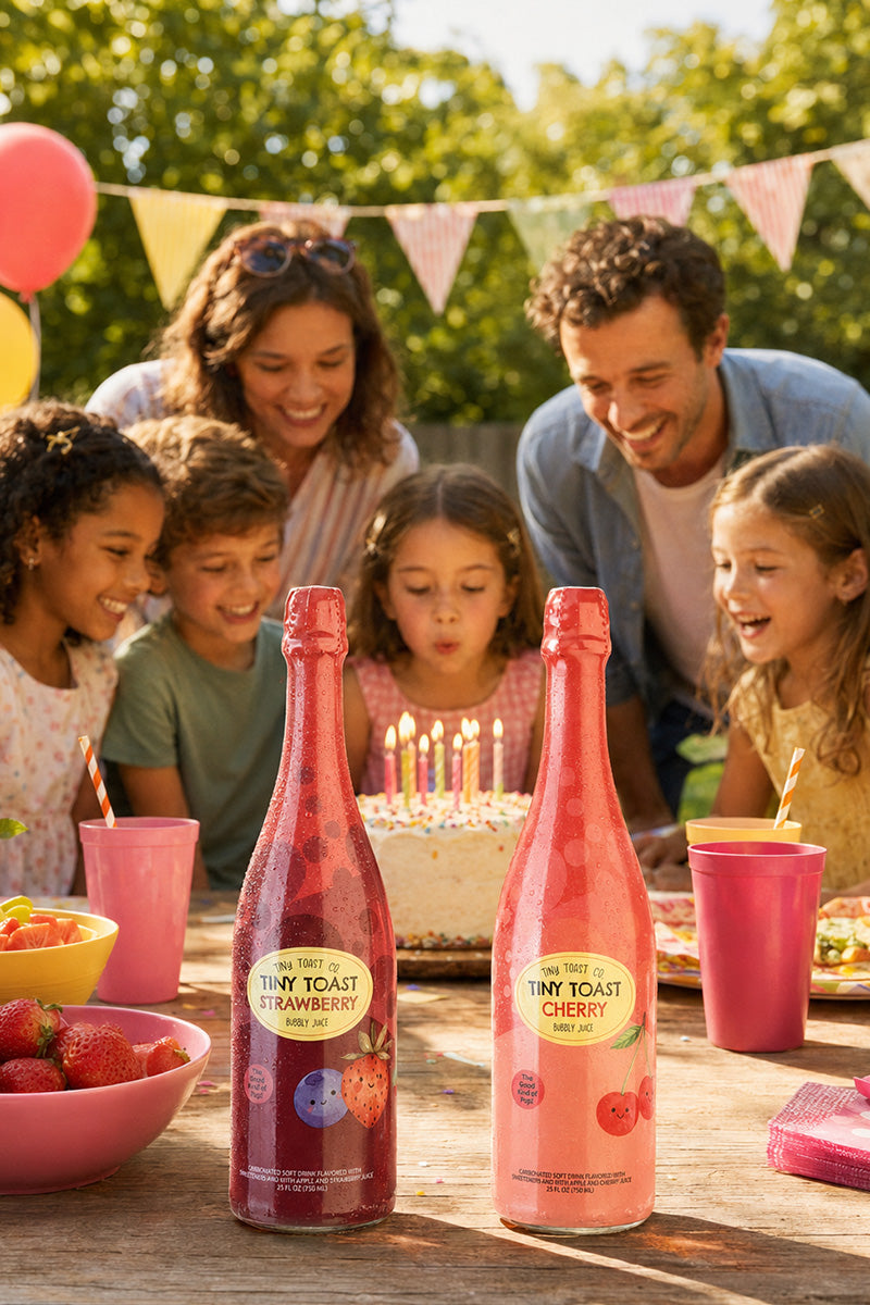 Two bottles of Tiny Toast sparkling drink, one Strawberry and one Cherry, displayed on a wooden table in front of a group of children and adults celebrating with a cake and balloons outdoors.
