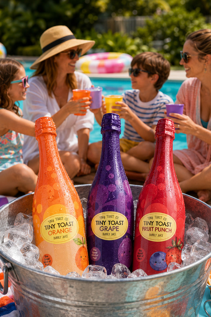 Three bottled fruit juices in colorful bottles arranged in a metal ice bucket, with a family sitting by a swimming pool in the background holding matching cups.