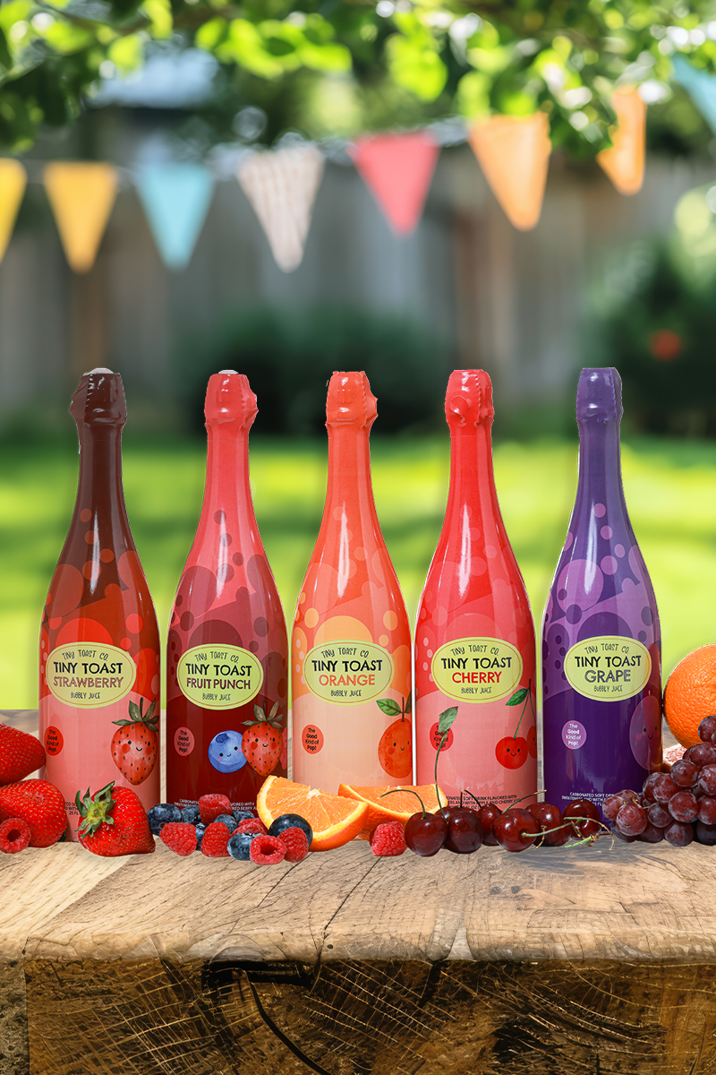 Five bottles of Tiny Toast sparkling fruit drink in strawberry, peach, orange, cherry, and grape flavors, lined up on a rustic wooden table with fresh fruit garnishes, set against a blurred outdoor garden background with colorful bunting.