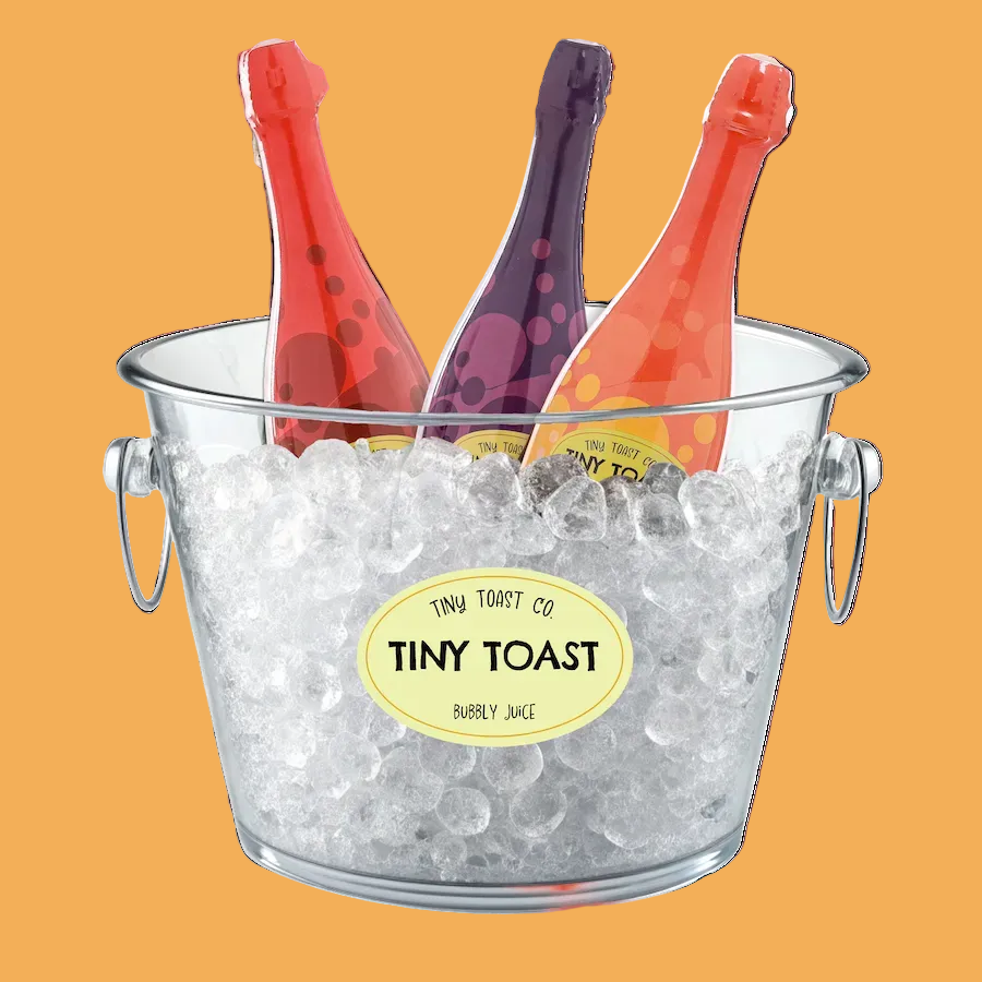A clear glass ice bucket filled with crushed ice, holding three miniature champagne bottles in red, purple, and orange, with a yellow oval label reading 'TINY TOAST CO. TINY TOAST BERRY JACE' on the front.